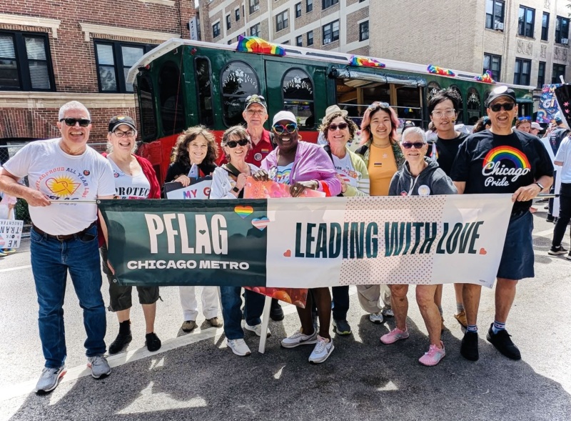 A group of 10 PFLAG Chicago Metro members holding a banner with a PFLAG decorated trolley in the background, at the Chicago Pride Parade.